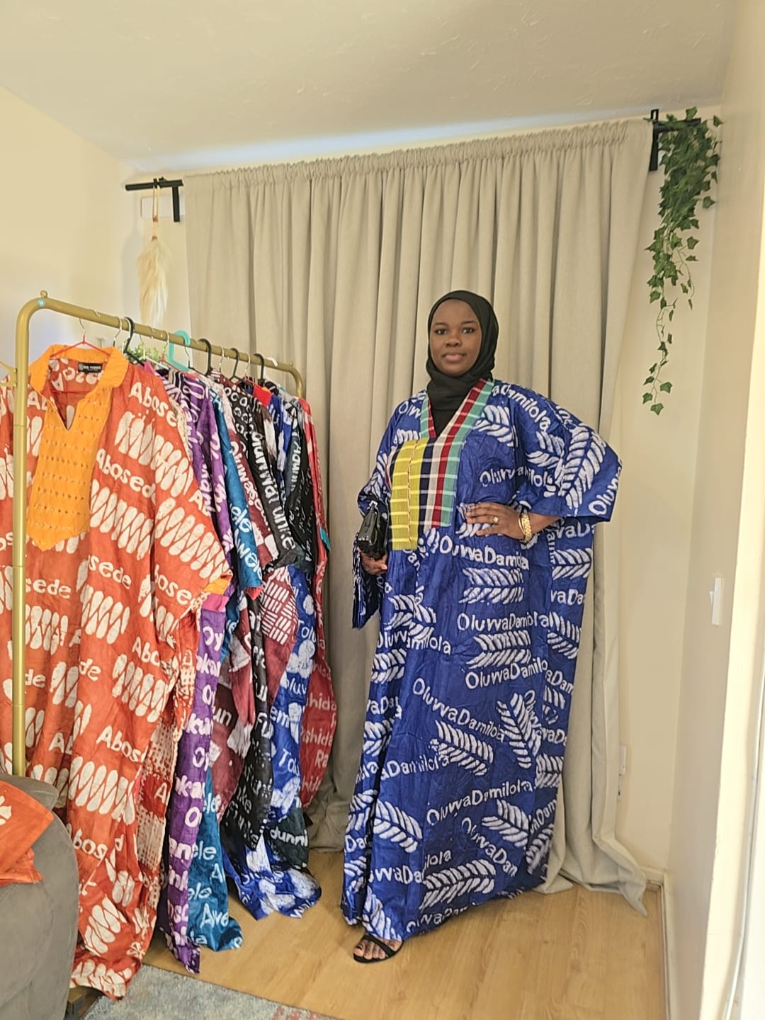 Woman standing next to a rack of colorful clothing in a room with curtains and a plant.